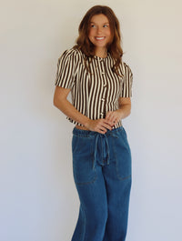 A woman wearing a brown and beige vertically striped short-sleeve button-up top with blue wide-leg jeans, standing against a plain light gray wall.