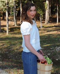 A woman with curly brown hair stands outdoors in a wooded area holding a wooden basket filled with green and red apples. She is wearing a white short-sleeve blouse with a ruffled collar and dark blue pants.