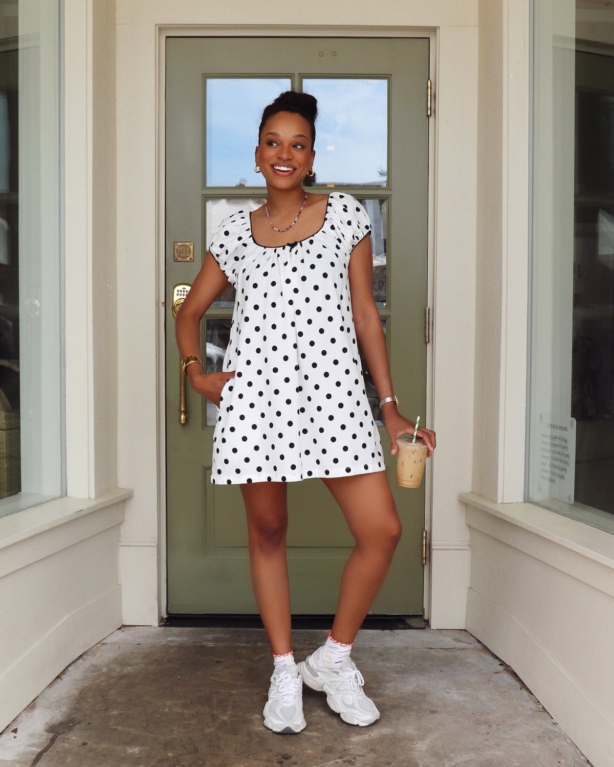 Woman in a polka dot dress standing in front of a door holding a drink.