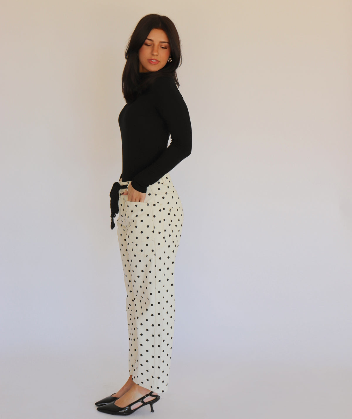 A woman in a black turtleneck and white barrel jeans with black polka dots on them, in front of a white background