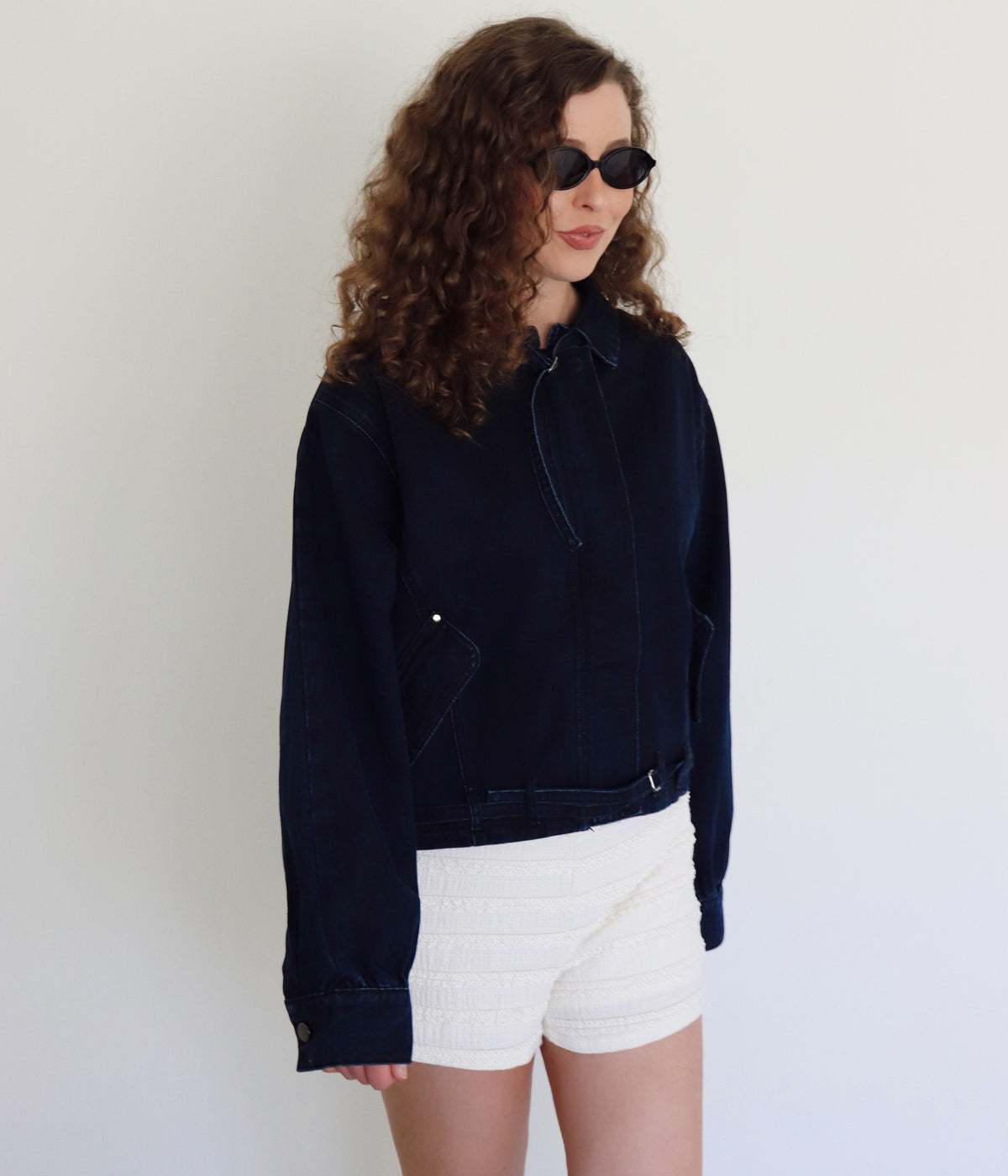 A woman with curly brown hair and sunglasses wearing a dark navy blue cropped denim jacket and white textured shorts, posing against a plain white wall.