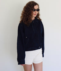 A woman with curly brown hair and sunglasses wearing a dark navy blue cropped denim jacket and white textured shorts, posing against a plain white wall.
