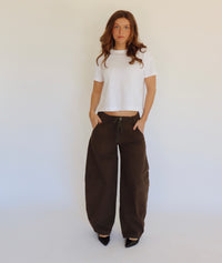 A woman stands against a plain white studio background wearing a white cropped short-sleeve top, loose dark brown wide-leg pants, and black pointed shoes.