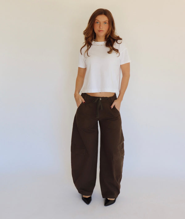 A woman stands against a plain white studio background wearing a white cropped short-sleeve top, loose dark brown wide-leg pants, and black pointed shoes.