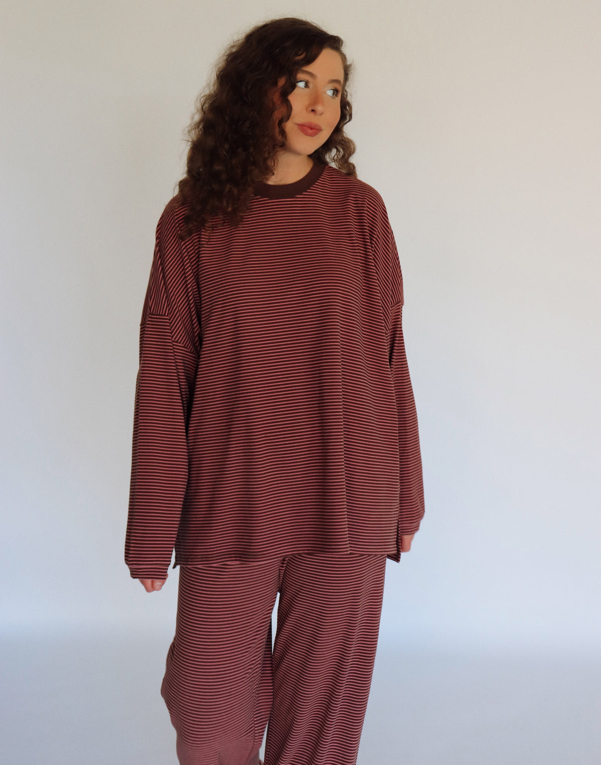 A woman modeling a matching two-piece loungewear set consisting of a long-sleeve top and pants, both featuring thin horizontal stripes in a pink and dark brown colorway.