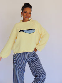 Person wearing a light yellow sweater with a fish design and striped pants against a plain background