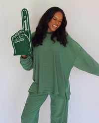 A woman wearing a green and yellow  striped long-sleeve top and matching pants, holding a green foam finger with white '#1' text, against a plain white background.