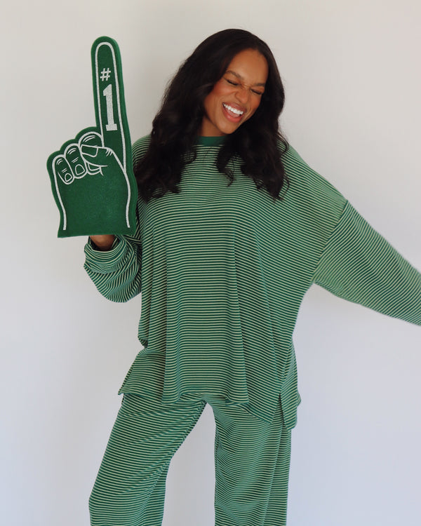 A woman wearing a green and yellow  striped long-sleeve top and matching pants, holding a green foam finger with white '#1' text, against a plain white background.
