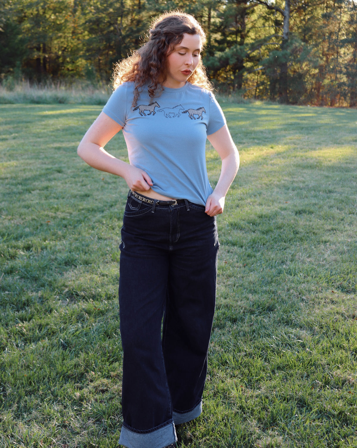A woman standing on a grassy lawn wearing a light blue graphic t-shirt and dark wide-leg jeans.