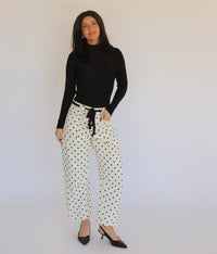 A woman in a black turtleneck and white barrel jeans with black polka dots on them, in front of a white background