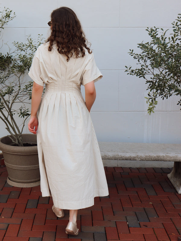 Woman in a taupe and cream striped  dress standing outdoors with plants and a bench in the background