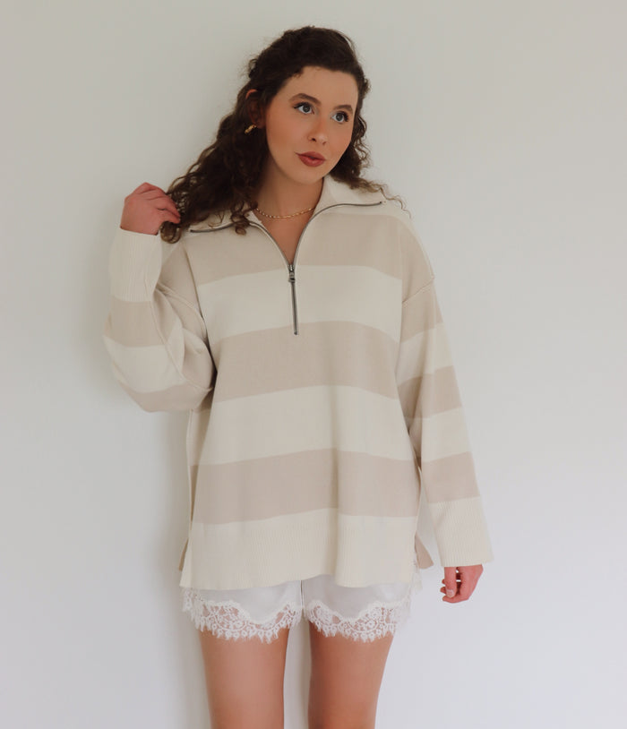 A woman wearing a cream and beige horizontally striped half-zip pullover sweater with white lace-trimmed shorts, standing against a plain white wall.