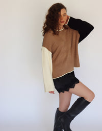 A woman modeling a color-block knit sweater with a brown body, black sleeves, and cream-colored underarms and cuffs, paired with black shorts and black knee-high boots against a plain white background.