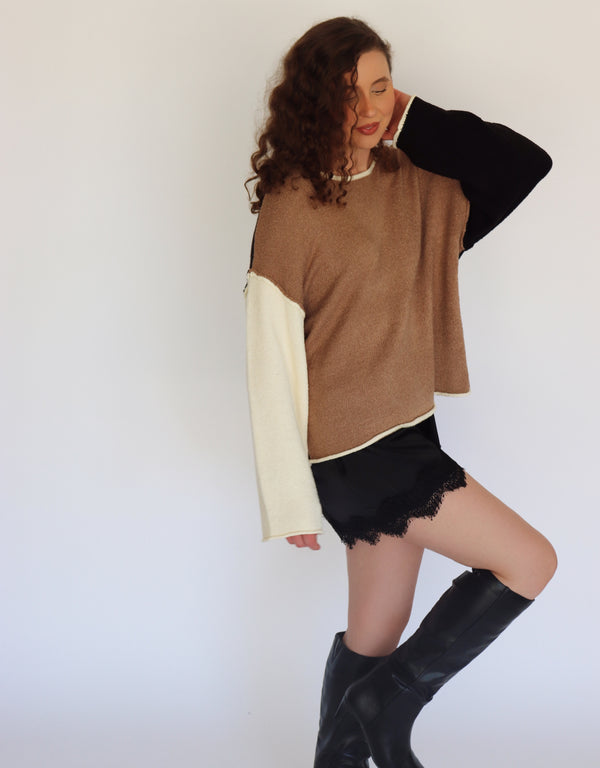 A woman modeling a color-block knit sweater with a brown body, black sleeves, and cream-colored underarms and cuffs, paired with black shorts and black knee-high boots against a plain white background.