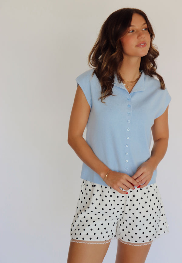 A woman modeling a light blue short-sleeve button-up shirt and white shorts with black polka dots, standing against a plain light background.