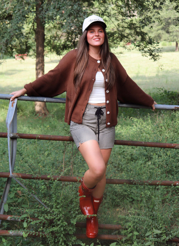 A woman posing outdoors on a rustic metal gate, wearing a brown knit cardigan, a white top, a white and black gingham shorts and red rain boots.