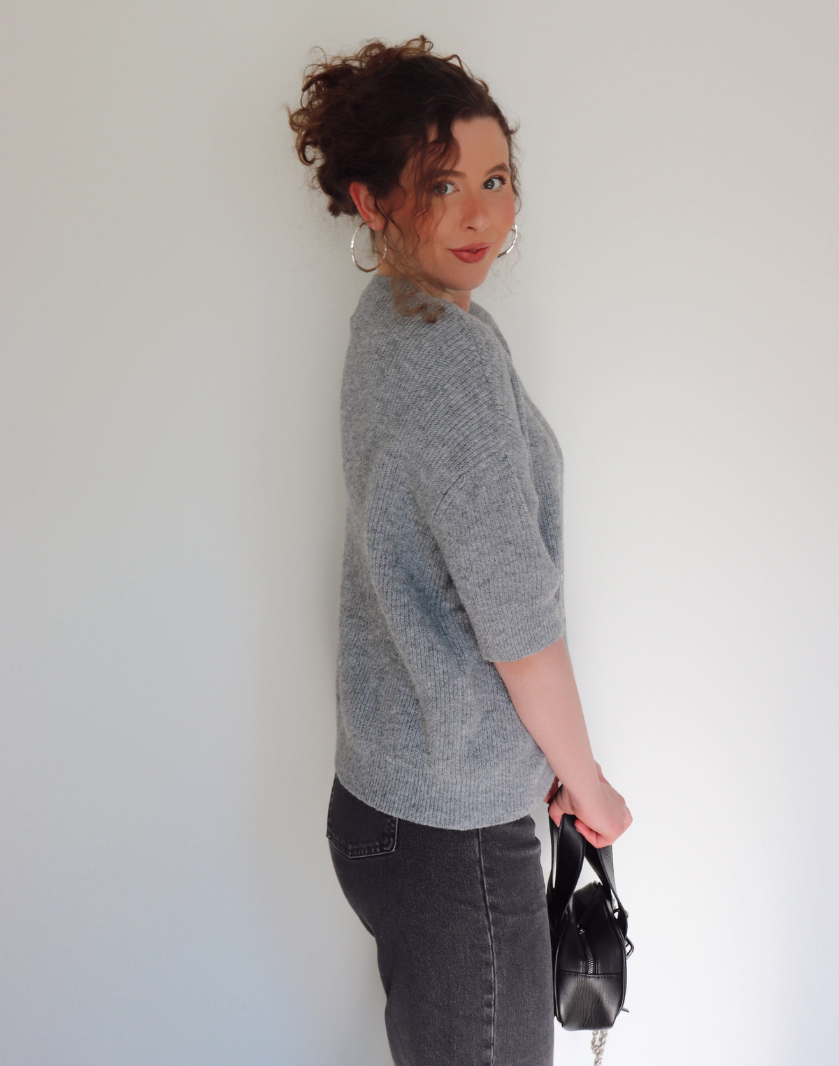Woman wearing a gray sweater and dark jeans, holding a black handbag against a white wall.