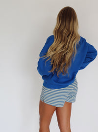 A woman modeling a royal blue mock neck sweatshirt and blue and white striped shorts against a plain white wall.