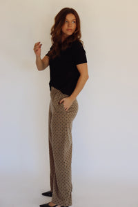 A person wearing wide-leg pants in a warm taupe-brown color with a small black polka dot pattern, paired with a black top and black heels, standing against a plain white background.