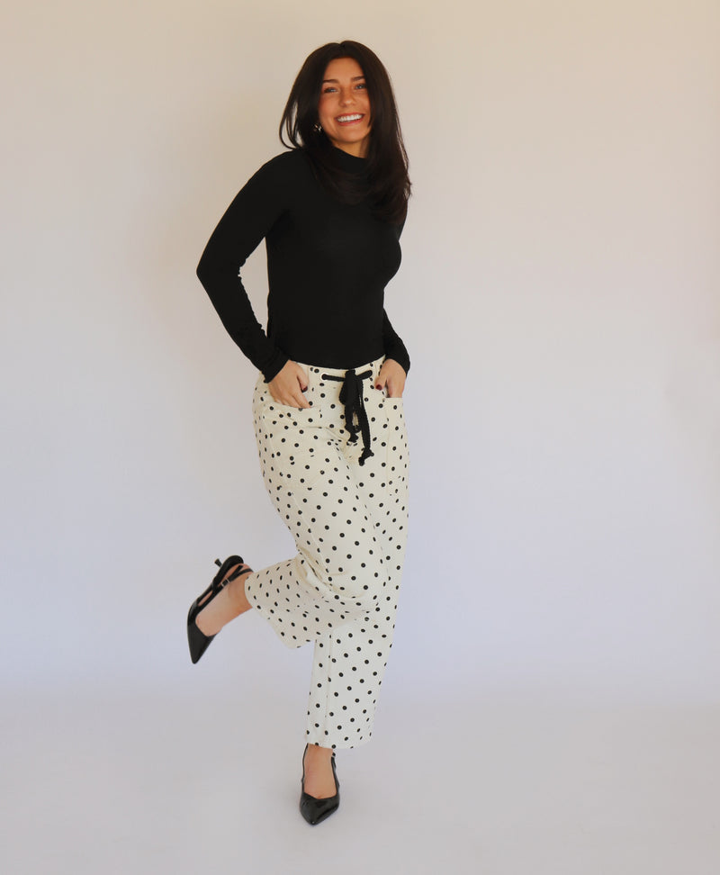 A woman in a black turtleneck and white barrel jeans with black polka dots on them, in front of a white background
