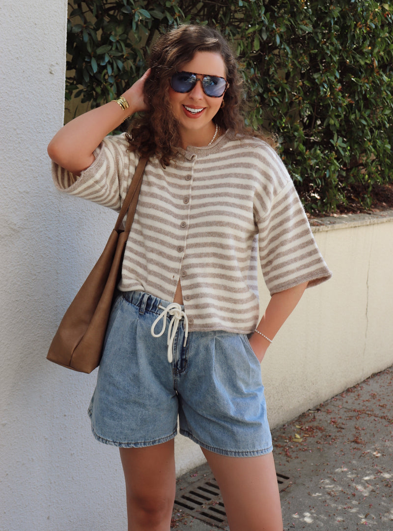 Woman wearing a striped cardigan and denim shorts with sunglasses, standing against a wall with greenery.