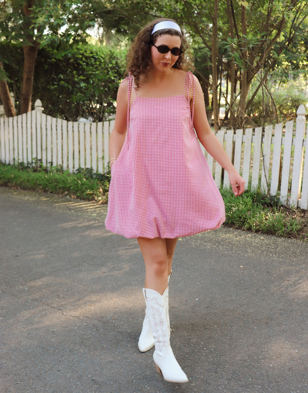 Woman in a red checkered dress and white boots standing on a path with trees and a white fence in the background.