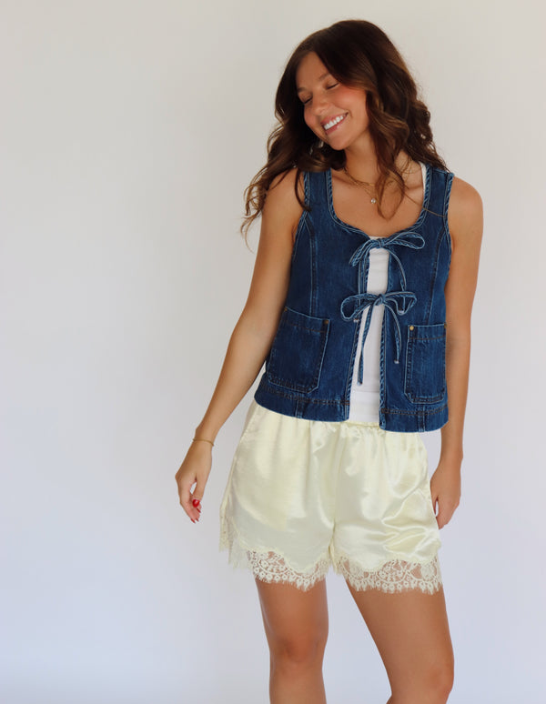A woman modeling a sleeveless blue denim vest with front tie details over a white top and yellow satin shorts with lace trim, set against a plain background.