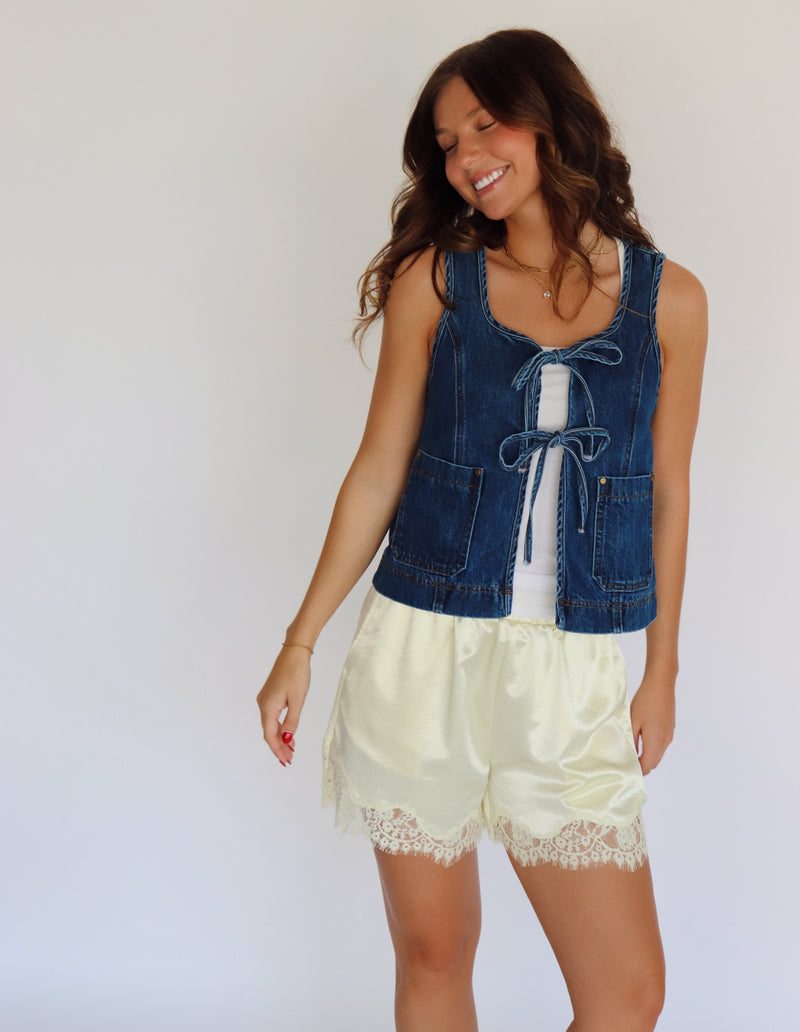 A woman modeling a sleeveless blue denim vest with front tie details over a white top and yellow satin shorts with lace trim, set against a plain background.