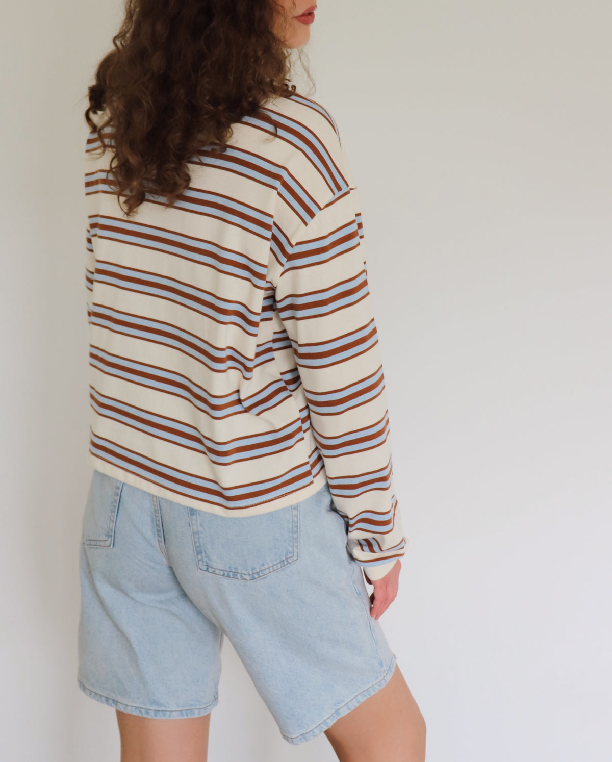 A woman shown from the back wearing a cream-colored long-sleeve knit top with blue and camel horizontal stripes, paired with light-wash denim shorts, against a plain white wall.