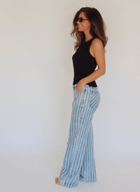 A woman wearing a black sleeveless top and light blue and white vertically striped wide-leg jeans, standing in profile against a plain white studio background.