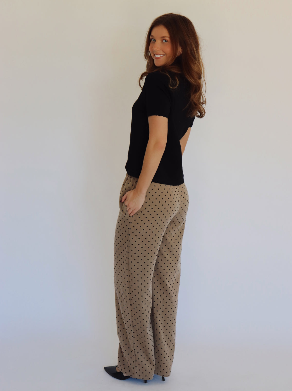 A person wearing wide-leg pants in a warm taupe-brown color with a small black polka dot pattern, paired with a black top and black heels, standing against a plain white background.