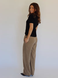 A person wearing wide-leg pants in a warm taupe-brown color with a small black polka dot pattern, paired with a black top and black heels, standing against a plain white background.