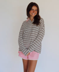 A woman modeling a brown, pink and white horizontally striped long-sleeve polo shirt with a pink collar, paired with light pink shorts, against a plain white background.