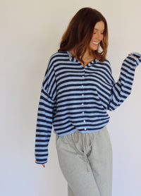 A woman wearing a navy and light blue horizontally striped long-sleeve button-up cardigan and light gray sweatpants, standing against a plain off-white wall.