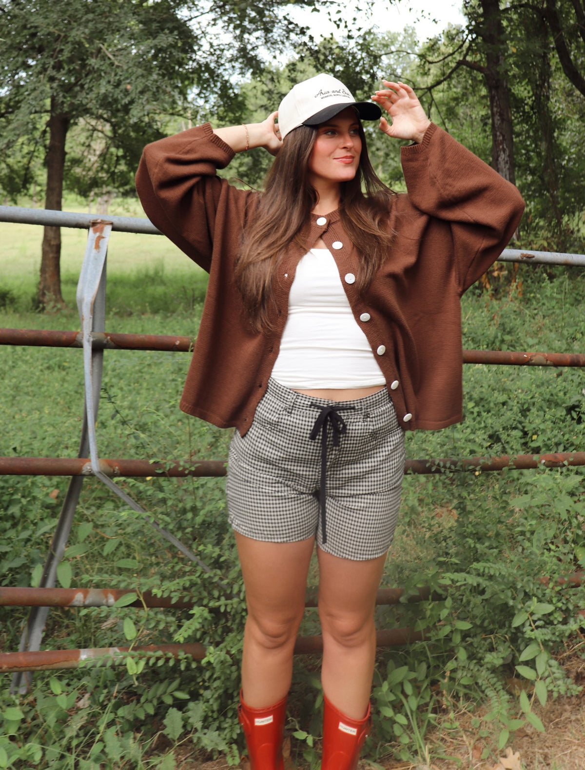 A woman posing outdoors in front of a metal fence and green trees, wearing a brown button-up cardigan, a white ribbed tank top, black and white gingham shorts, red rain boots, and a white baseball cap.