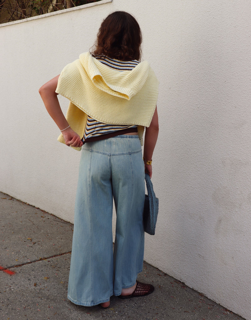 Woman wearing a striped shirt, a yellow sweater around her shoulders  and blue jeans against a gray wall.