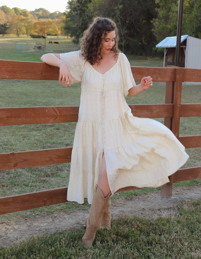 Woman in a cream dress and beige boots standing near a wooden fence in an outdoor setting.