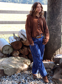 A woman stands outdoors next to a tree and a wooden fence, wearing a brown button-up cardigan and blue striped pants.