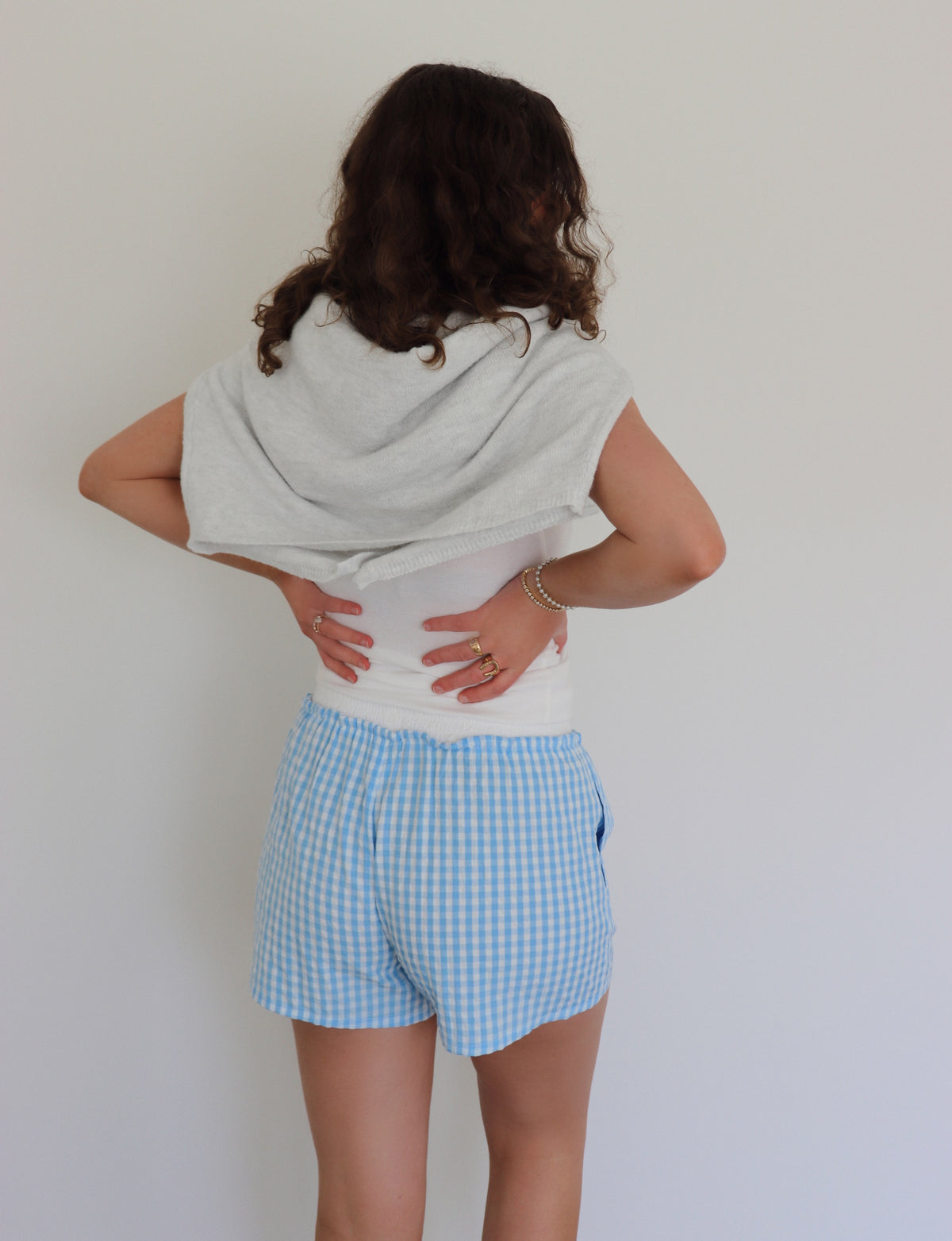 Woman wearing a white tank top, a light gray cardigan tied around her shoulders and blue checkered shorts against a white background