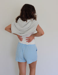 Woman wearing a white tank top, a light gray cardigan tied around her shoulders and blue checkered shorts against a white background