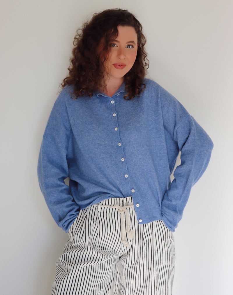 Woman wearing a blue cardigan and striped pants in front of a white wall.