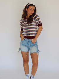 A woman modeling a short-sleeve knit polo shirt with horizontal stripes in cream, burgundy, and blue, paired with light-wash denim shorts.