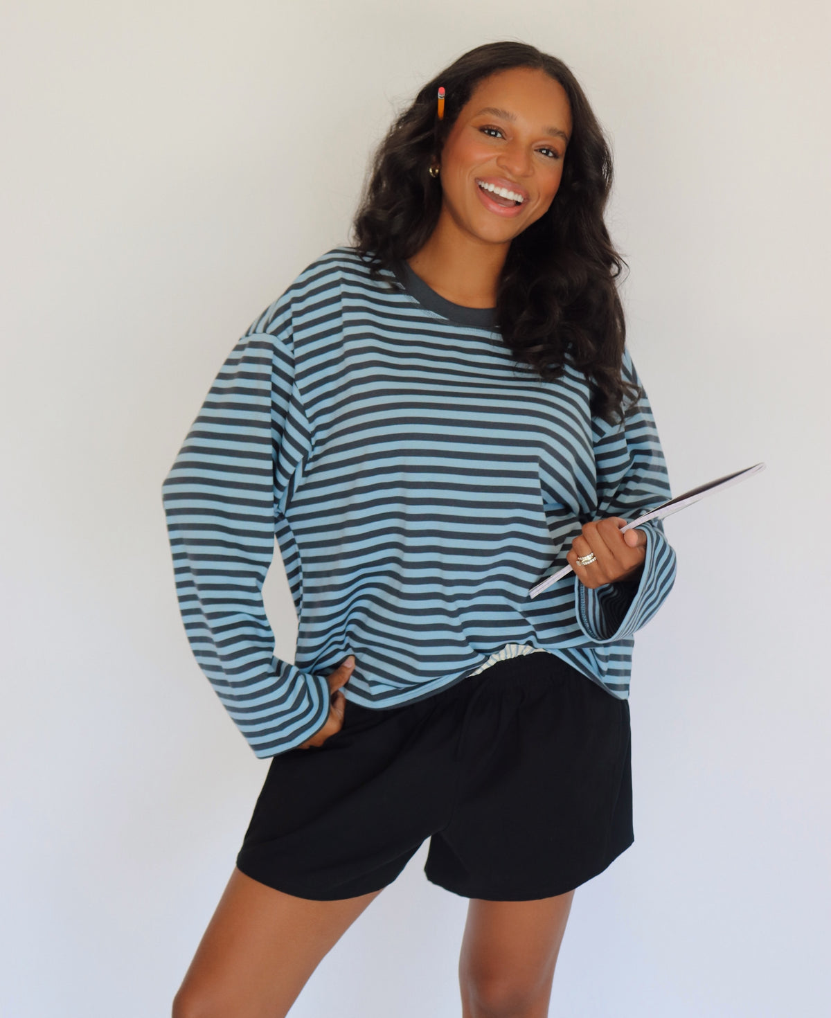 A woman modeling a long-sleeve blue striped top with shorts, holding a black and white notebook bag against a plain white background.