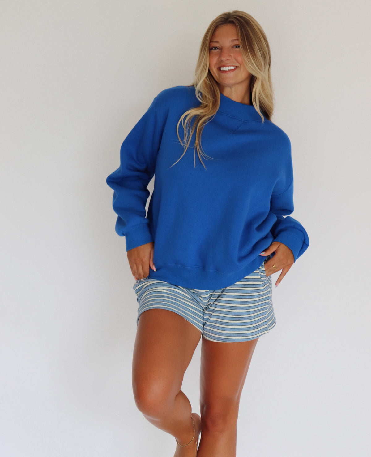 A woman modeling a royal blue mock neck sweatshirt and blue and white striped shorts against a plain white wall.