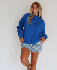 A woman modeling a royal blue mock neck sweatshirt and blue and white striped shorts against a plain white wall.