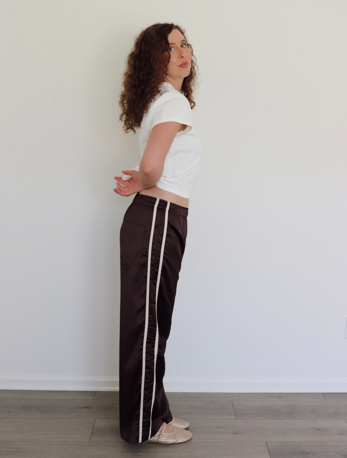 A woman with curly brown hair stands in profile wearing dark brown wide-leg pants with a white stripe down the side, a white cropped t-shirt, and beige flats, set against a plain white wall and light wood floor.
