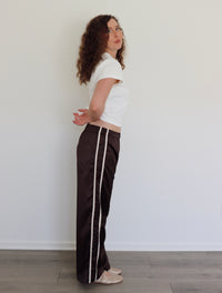 A woman with curly brown hair stands in profile wearing dark brown wide-leg pants with a white stripe down the side, a white cropped t-shirt, and beige flats, set against a plain white wall and light wood floor.