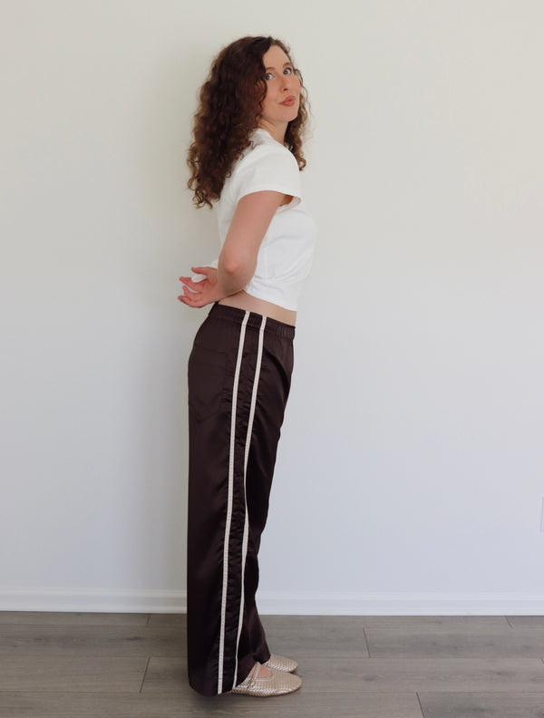 A woman with curly brown hair stands in profile wearing dark brown wide-leg pants with a white stripe down the side, a white cropped t-shirt, and beige flats, set against a plain white wall and light wood floor.