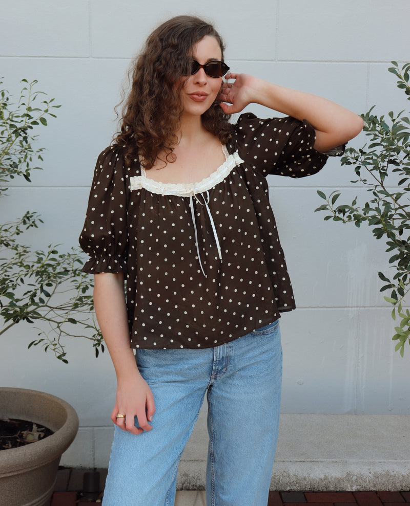 Woman wearing a brown polka dot blouse and blue jeans standing outdoors with plants in the background.