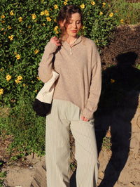 Person outdoors with greenery and flowers in the background wearing a brown top and striped pants.